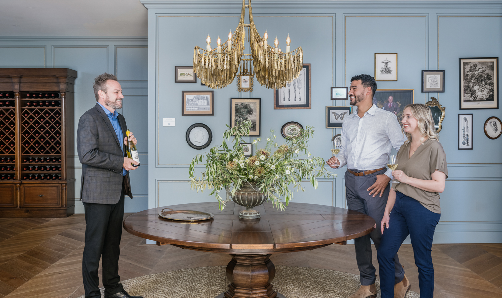 group of people holding glasses of jordan chardonnay standing by round wooden table decorated with floral arrangement underneath brass chandelier