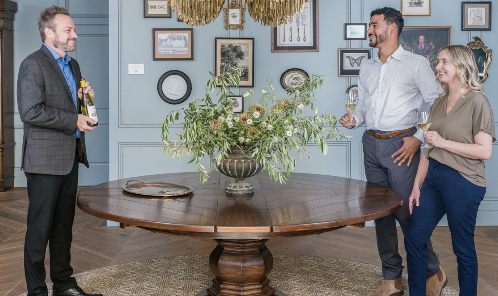 group of people holding glasses of jordan chardonnay standing by round wooden table decorated with floral arrangement underneath brass chandelier