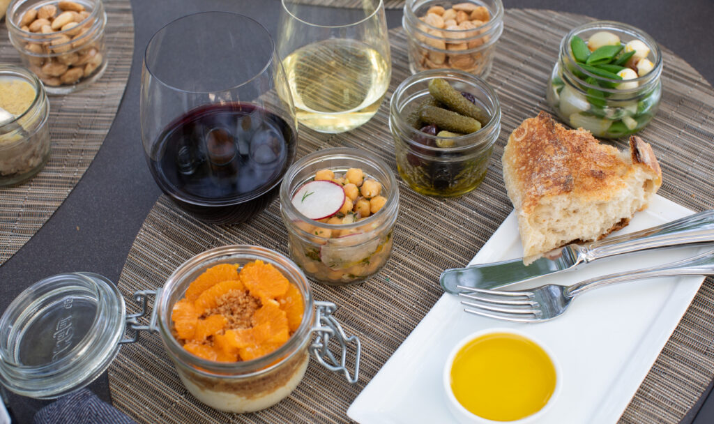 Picnic culinary bites in presented in glass jars with stemless glasses of Cabernet Sauvignon and Chardonnay on an outdoor table