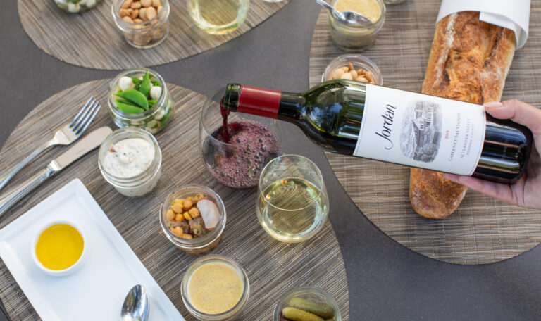 Overhead image of Jordan Cabernet Sauvignon being poured into stemless wine glass with a baguette and other picnic bites on an outdoor table