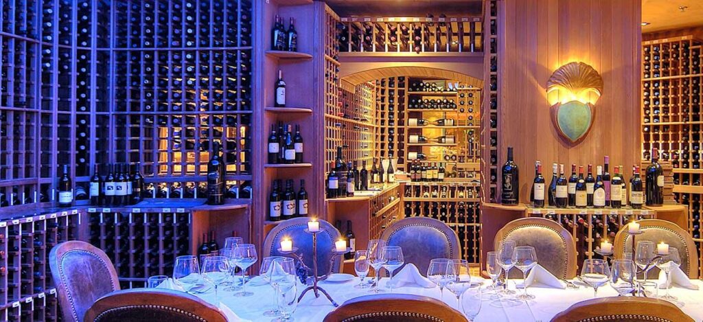 A restaurant table set for dinner with a white table cloth and numerous wine glasses, surrounded by cellar shelves filled with wine bottles