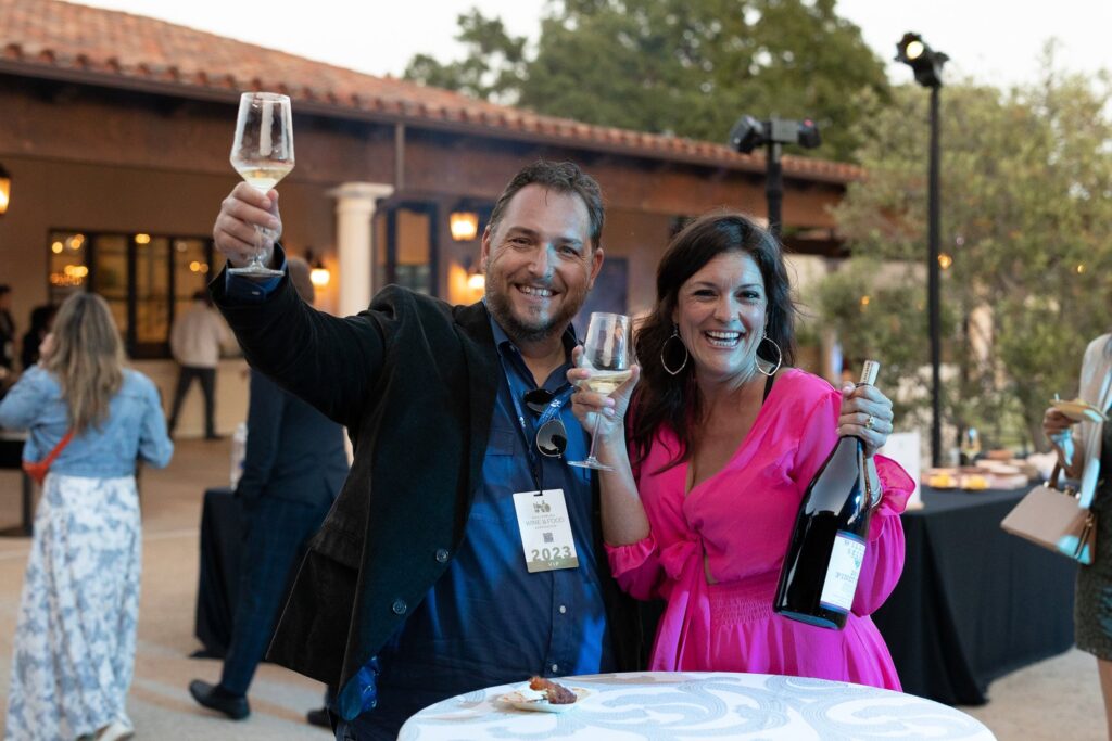 a couple posing with raised wine glasses and a magnum bottle of pinot noir